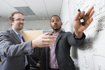 Two faculty members pointing to a whiteboard as they discuss something.