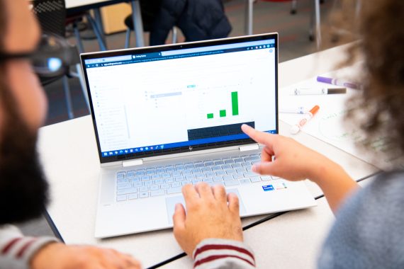 Two people looking at a laptop screen. The person on the right points to a bar graph on the screen.