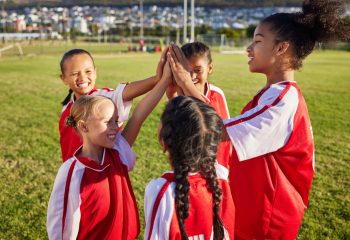 Children in soccer uniforms bring their hands together in a group high five on a soccer field.
