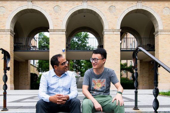 A professor and Ph.D. student sit on a step, talking together, outside on the Medford/Somerville campus