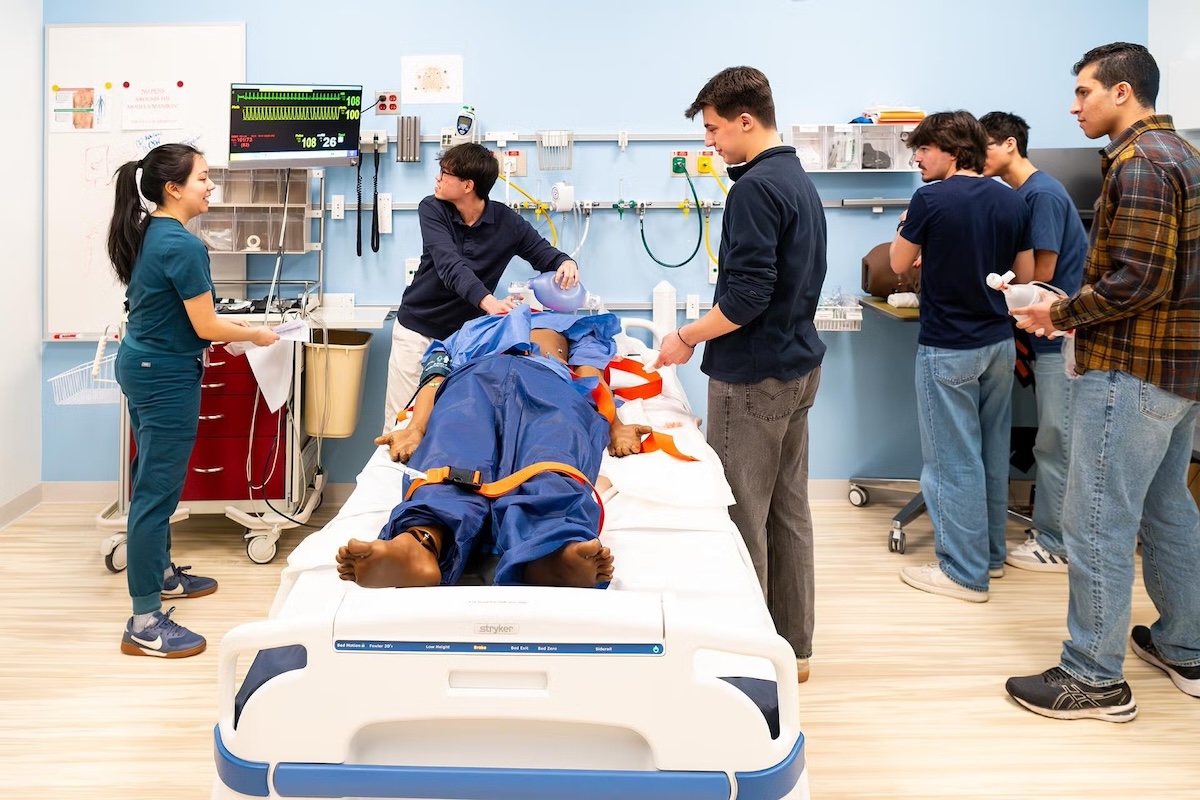A Tufts medical student wearing scrubs coaching five Tufts undergraduate students as they respond to a simulated emergency on a mannequin in the Tufts University School of Medicine Simulation Center.