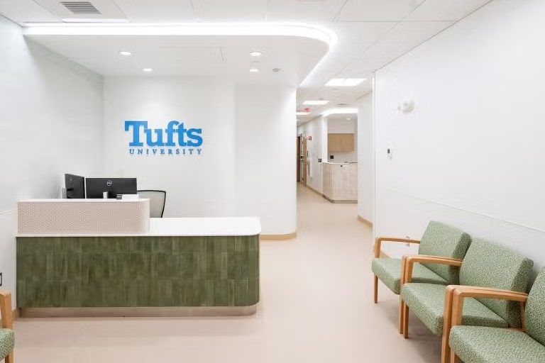 Image of the waiting area in the new student health clinic. The Tufts University logo is seen on the wall behind the front desk. There are chairs in the waiting area and a hallway leading back into the facility.